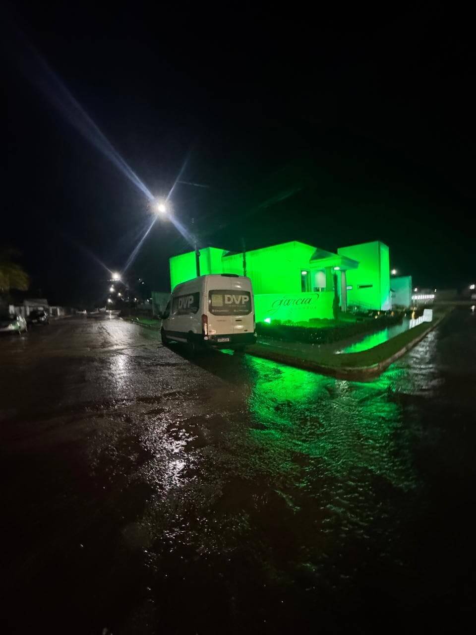 Iluminação cênica arquitetural em verde destacando a fachada de um prédio à noite, com van da DVP Som e Luz estacionada.