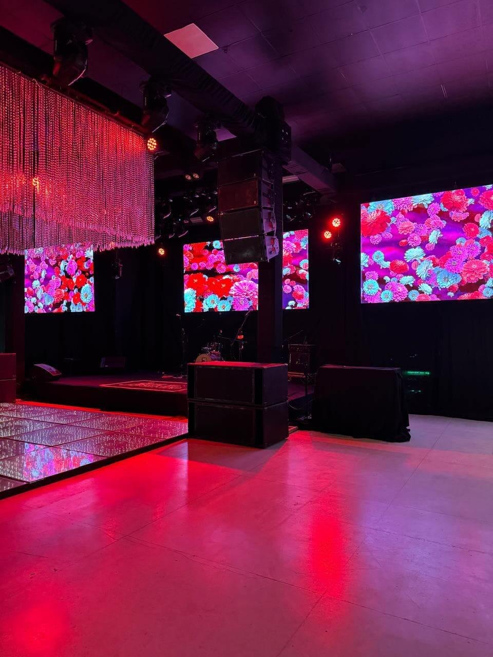 Salão de festas com pista de dança iluminada, painéis de LED ao fundo com projeções coloridas e estrutura de teto com cristais e luzes vermelhas.
