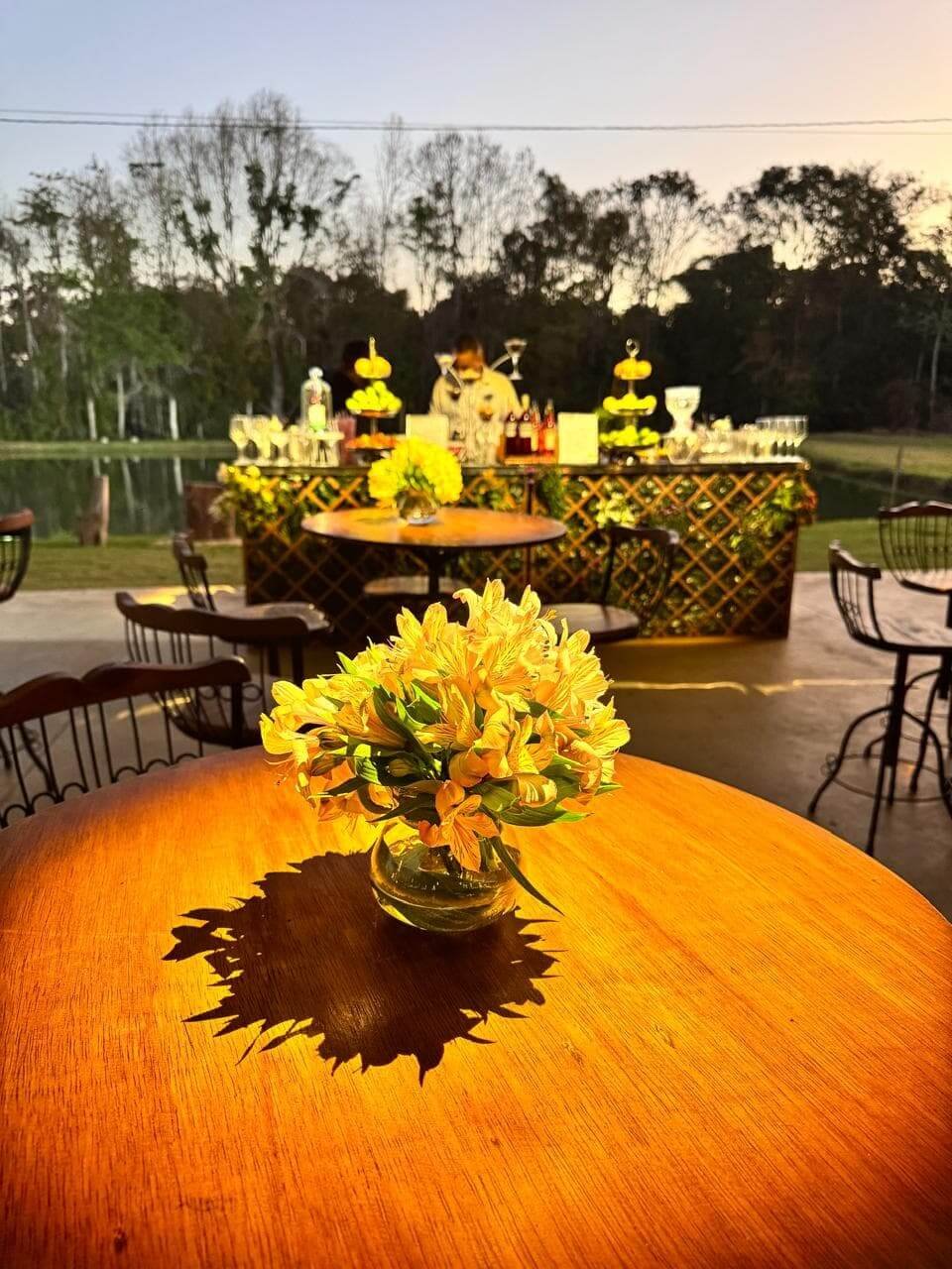 Detalhe de mesa decorada com arranjo de flores amarelas e iluminação quente em área externa de evento com vista para lago.
