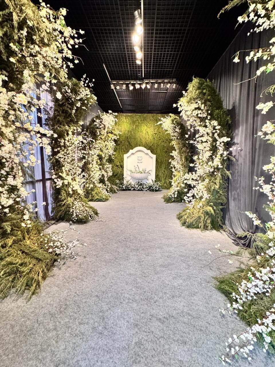 Passarela de entrada de evento decorada com arcos de flores brancas e folhagens, iluminada por luzes embutidas no teto e nas laterais.