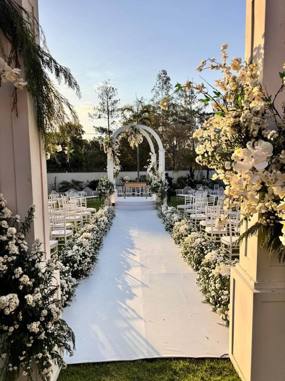 Cerimônia de casamento clássica ao ar livre com passarela de carpete branco, cadeiras brancas e arco floral ao fundo.