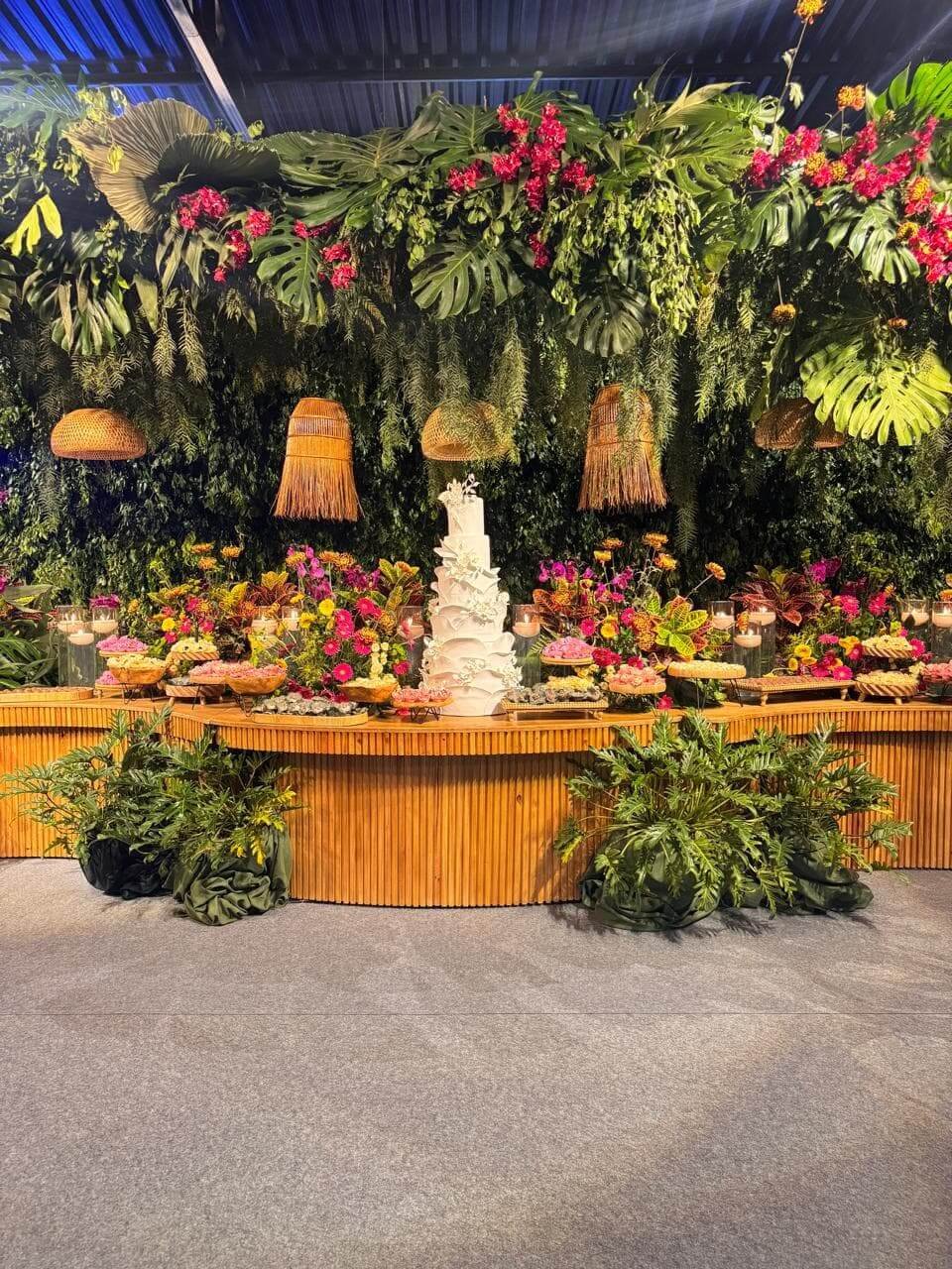 Mesa de bolo e doces com decoração tropical exuberante, parede verde de folhagens e luminárias rústicas pendentes.