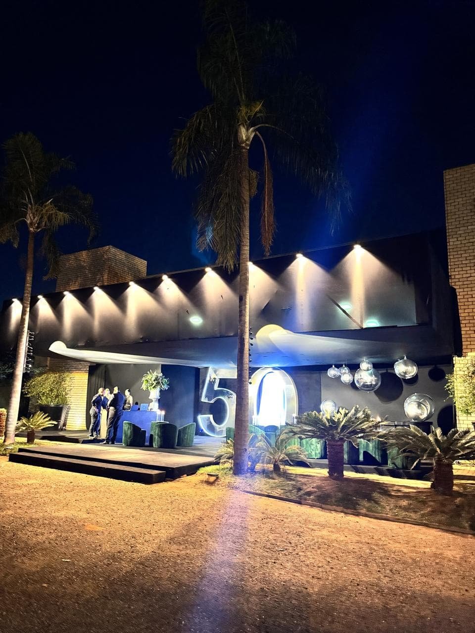 Fachada de local de evento iluminada à noite, com destaque para o paisagismo e letreiro comemorativo de 50 anos.