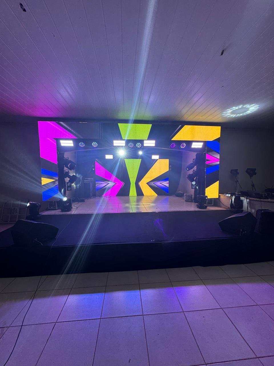 Palco com grande painel de LED exibindo gráficos geométricos coloridos e feixes de luz moving head cortando o palco.