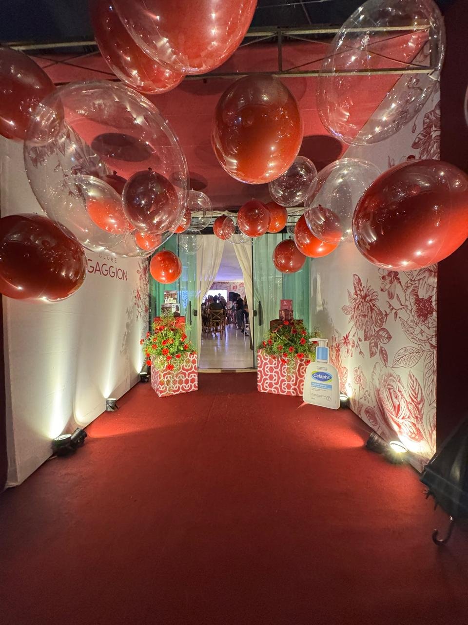 Entrada de evento decorada com tapete vermelho e teto coberto por balões vermelhos e transparentes criando um túnel visual.