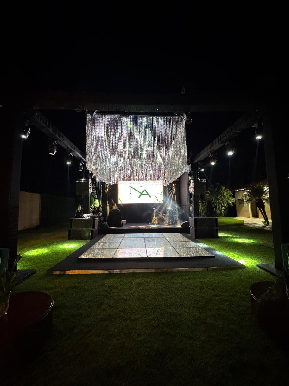 Estrutura de palco e pista de dança montada em gramado ao ar livre, com cortina de cristais e iluminação noturna.