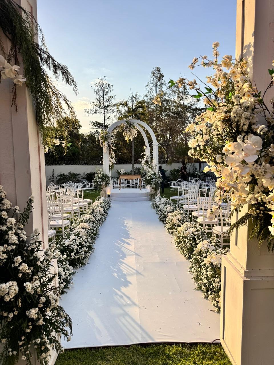 Cenário de casamento clássico ao ar livre com passarela branca, cadeiras Tiffany brancas e arcos de flores na entrada.