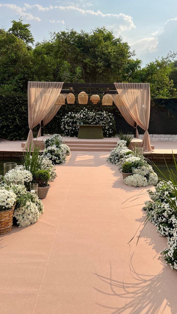 Altar de casamento estilo boho chic ao ar livre, com passarela bege, arranjos de flores brancas e pergola com luminárias de palha.