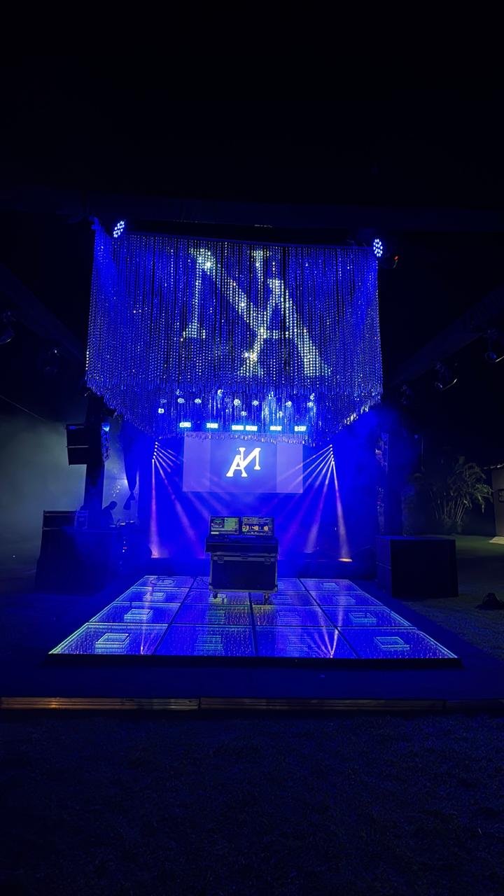 Palco de DJ iluminado em azul com piso de LED, cortina de cristais e projeção de monograma neon personalizado.
