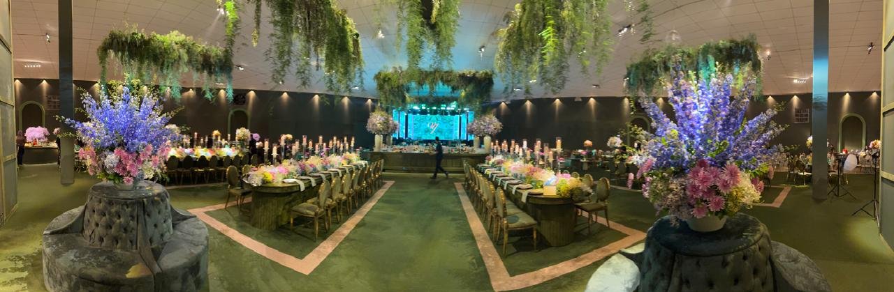 Salão de banquetes sofisticado com mesas longas, decoração aérea com folhagens verdes e palco ao fundo.