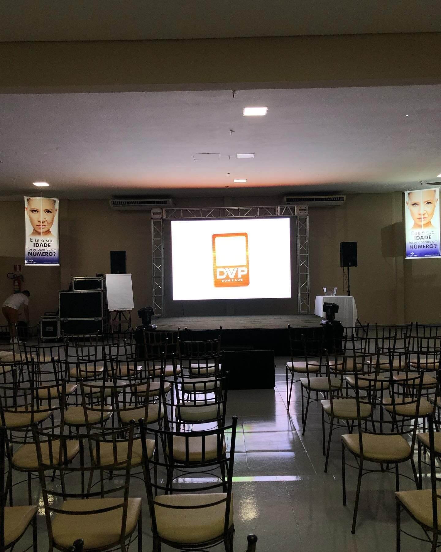 Auditório para evento corporativo organizado com fileiras de cadeiras Tiffany douradas e palco equipado com telão de projeção para apresentações e palestras.
