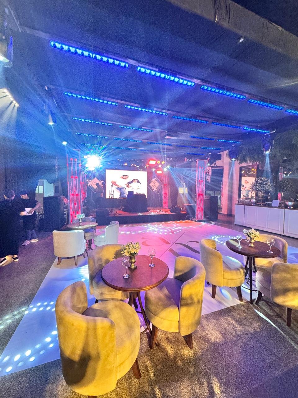 Lounge de evento social com iluminação ambiente azul e branca, mobiliário sofisticado e vista para a área de dança.